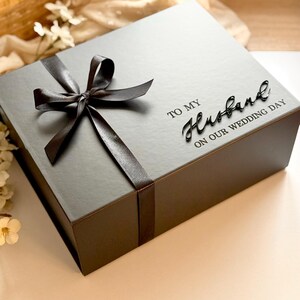 Puede incluir: Una caja de regalo gris atada con una cinta y lazo de sat&eacute;n marr&oacute;n oscuro. La caja tiene las palabras "TO MY Husband ON OUR WEDDING DAY" en una fuente de escritura. La caja est&aacute; sobre una superficie blanca.