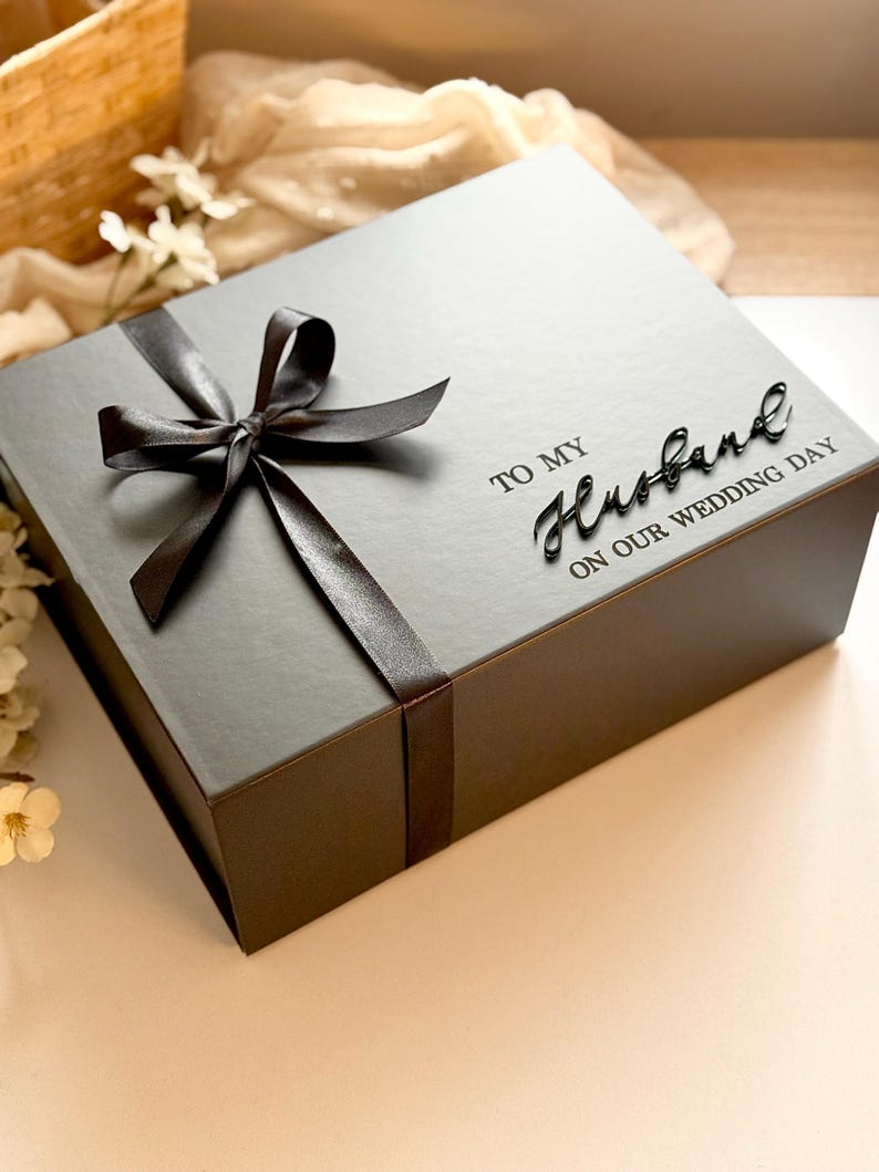 Puede incluir: Una caja de regalo gris atada con una cinta y lazo de sat&eacute;n marr&oacute;n oscuro. La caja tiene las palabras "TO MY Husband ON OUR WEDDING DAY" en una fuente de escritura. La caja est&aacute; sobre una superficie blanca.