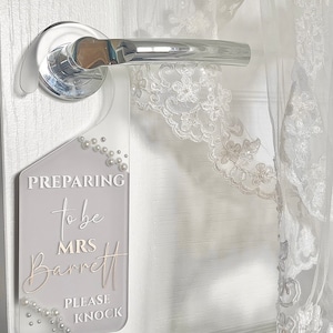 Puede incluir: Un colgador de puerta de acr&iacute;lico transparente con el texto "PREPARING to be MRS Barrett PLEASE KNOCK" en blanco y dorado. El colgador est&aacute; decorado con perlas de imitaci&oacute;n y cuelga de un tirador de puerta cromado. Una prenda de encaje blanco es parcialmente visible.