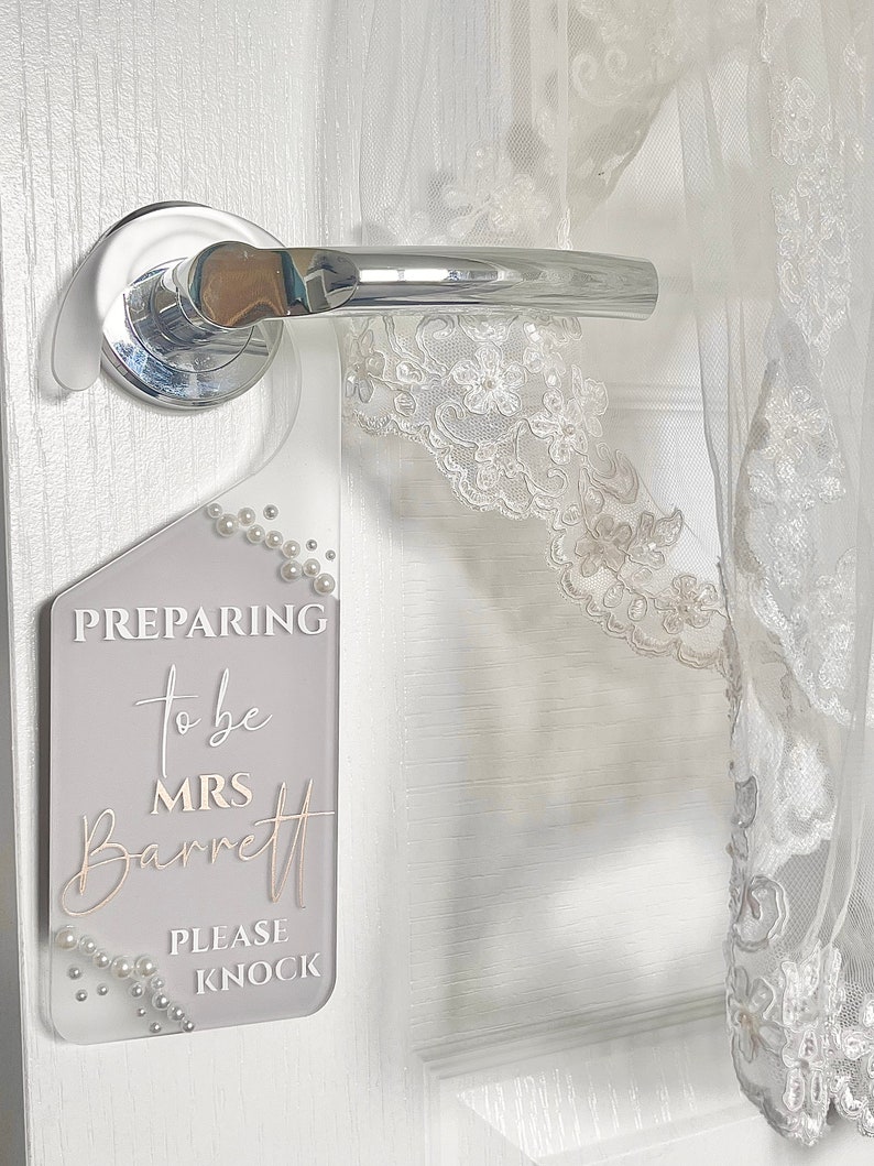 Puede incluir: Un colgador de puerta de acr&iacute;lico transparente con el texto "PREPARING to be MRS Barrett PLEASE KNOCK" en blanco y dorado. El colgador est&aacute; decorado con perlas de imitaci&oacute;n y cuelga de un tirador de puerta cromado. Una prenda de encaje blanco es parcialmente visible.