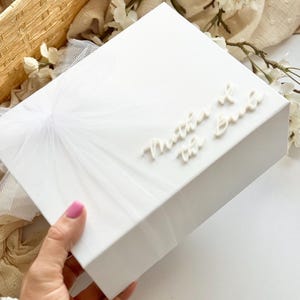 K&ouml;nnte beinhalten: Eine wei&szlig;e Geschenkbox mit dem Schriftzug "Mother of the Bride" in eleganter Schrift. Die Box hat eine strukturierte Oberfl&auml;che und ist mit einem zarten T&uuml;llmuster verziert. Der Hintergrund umfasst wei&szlig;e Blumen und einen Weidenkorb.