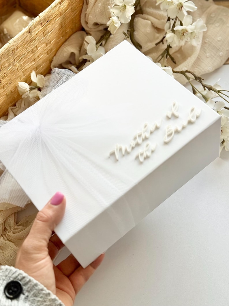 K&ouml;nnte beinhalten: Eine wei&szlig;e Geschenkbox mit dem Schriftzug "Mother of the Bride" in eleganter Schrift. Die Box hat eine strukturierte Oberfl&auml;che und ist mit einem zarten T&uuml;llmuster verziert. Der Hintergrund umfasst wei&szlig;e Blumen und einen Weidenkorb.
