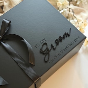 May include: A dark grey gift box tied with a black satin ribbon. The box is embossed with the words "TO MY Groom ON OUR WEDDING DAY" in a script font. A wedding gift box.