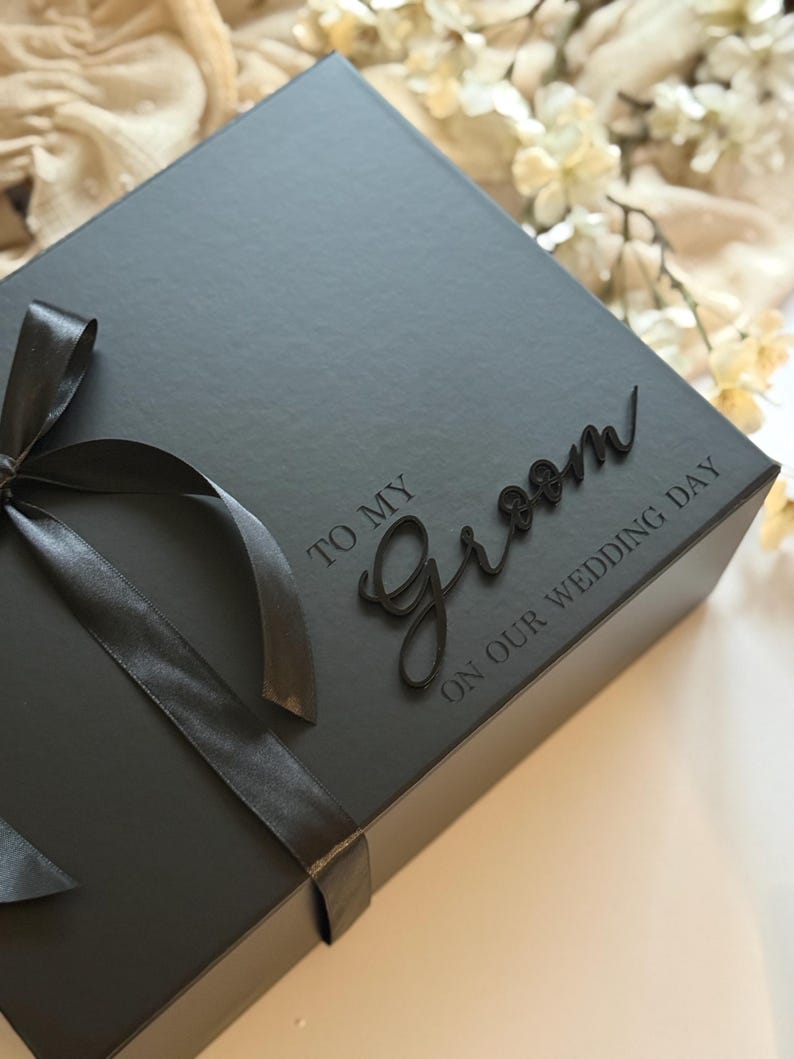May include: A dark grey gift box tied with a black satin ribbon. The box is embossed with the words "TO MY Groom ON OUR WEDDING DAY" in a script font. A wedding gift box.