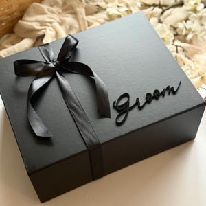 May include: A black gift box tied with a black satin ribbon and bow. The word "Groom" is written in cursive on the box. The box is on a white surface with a basket and flowers in the background.