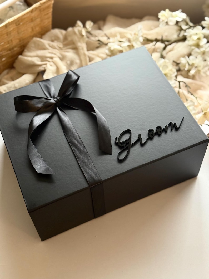 May include: A black gift box tied with a black satin ribbon and bow. The word "Groom" is written in cursive on the box. The box is on a white surface with a basket and flowers in the background.