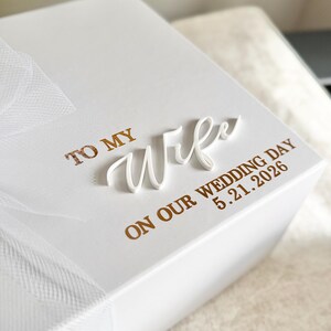 May include: A white gift box with the words "TO MY Wife" in gold lettering and a white cursive "Wife" overlay. The text "ON OUR WEDDING DAY 5.21.2026" is also in gold. A white tulle ribbon is tied around the box.