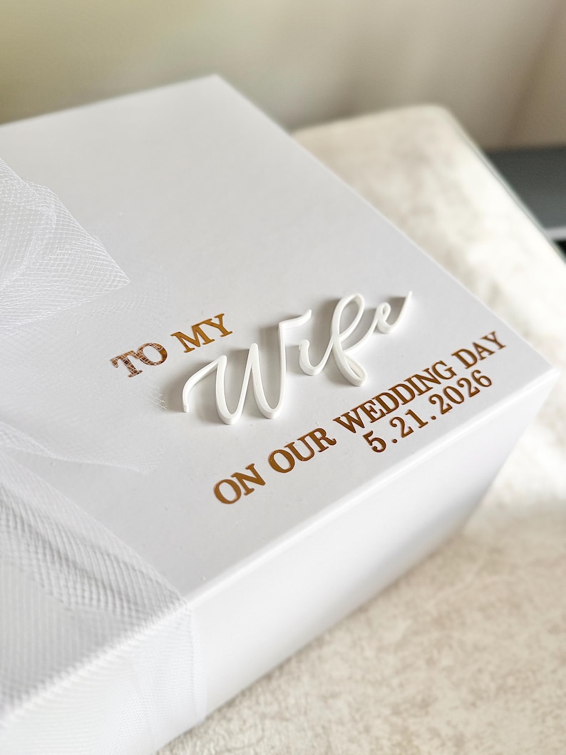 May include: A white gift box with the words "TO MY Wife" in gold lettering and a white cursive "Wife" overlay. The text "ON OUR WEDDING DAY 5.21.2026" is also in gold. A white tulle ribbon is tied around the box.