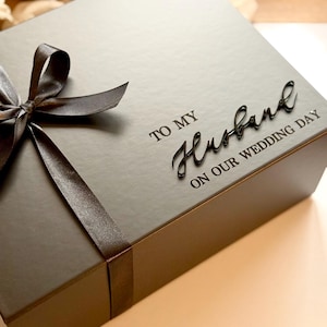 Puede incluir: Una caja de regalo gris oscuro atada con una cinta y lazo de sat&eacute;n marr&oacute;n. La caja est&aacute; inscrita con las palabras "TO MY Husband ON OUR WEDDING DAY" en una fuente elegante, ideal para un regalo de boda.