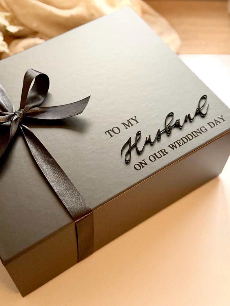 Puede incluir: Una caja de regalo gris oscuro atada con una cinta y lazo de sat&eacute;n marr&oacute;n. La caja est&aacute; inscrita con las palabras "TO MY Husband ON OUR WEDDING DAY" en una fuente elegante, ideal para un regalo de boda.