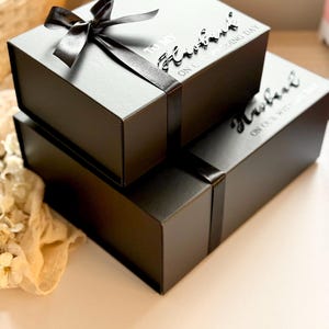Puede incluir: Dos cajas de regalo negras apiladas, atadas con cinta de raso negro. La caja superior tiene un lazo. El texto en las cajas dice "TO MY Husband ON OUR WEDDING DAY".