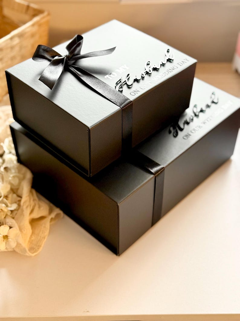 Puede incluir: Dos cajas de regalo negras apiladas, atadas con cinta de raso negro. La caja superior tiene un lazo. El texto en las cajas dice "TO MY Husband ON OUR WEDDING DAY".