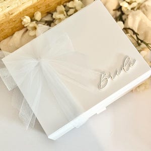 May include: A white gift box with a sheer white bow and the word "Bride" in cursive script. The box is rectangular and has a clean, minimalist design, perfect for a wedding or bridal shower.