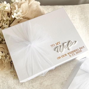 May include: A white gift box with a tulle bow and the inscription "TO MY Wife ON OUR WEDDING DAY 5.21.2026" in gold lettering. The box is on a textured surface, with white flowers in the background.