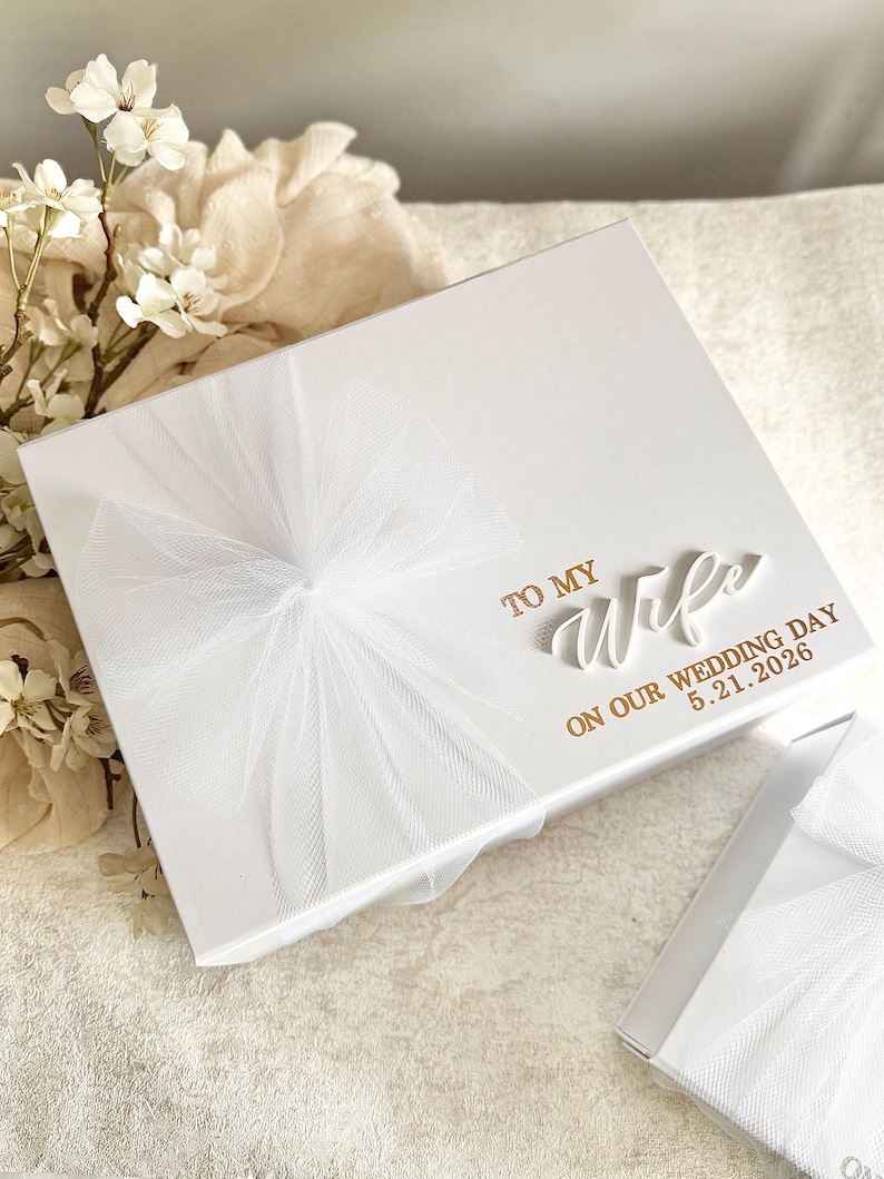 May include: A white gift box with a tulle bow and the inscription "TO MY Wife ON OUR WEDDING DAY 5.21.2026" in gold lettering. The box is on a textured surface, with white flowers in the background.