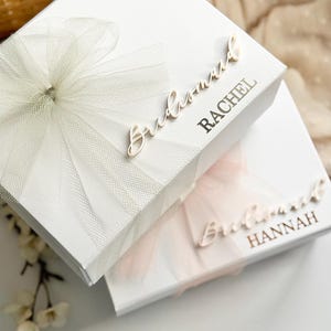 May include: Two white gift boxes, one with a white tulle bow and the name "Rachel" in gold lettering, and the other with a pink tulle bow and the name "Hannah". The word "Bridesmaid" is written in cursive on both boxes.