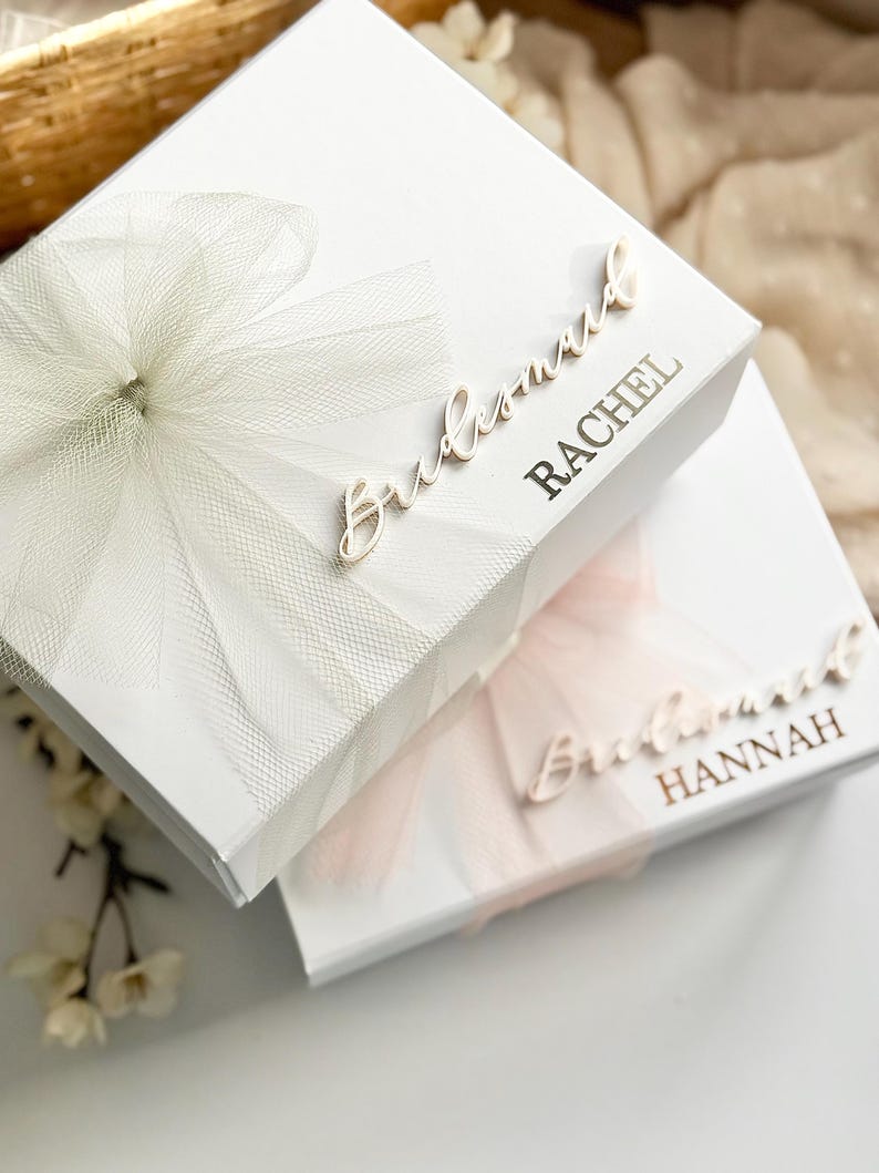 May include: Two white gift boxes, one with a white tulle bow and the name "Rachel" in gold lettering, and the other with a pink tulle bow and the name "Hannah". The word "Bridesmaid" is written in cursive on both boxes.