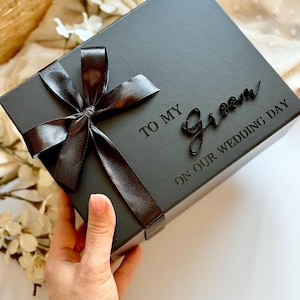 May include: A black gift box tied with a dark brown satin ribbon bow. The box is inscribed with the words "TO MY Groom ON OUR WEDDING DAY" in a script font. The box is held by a person.