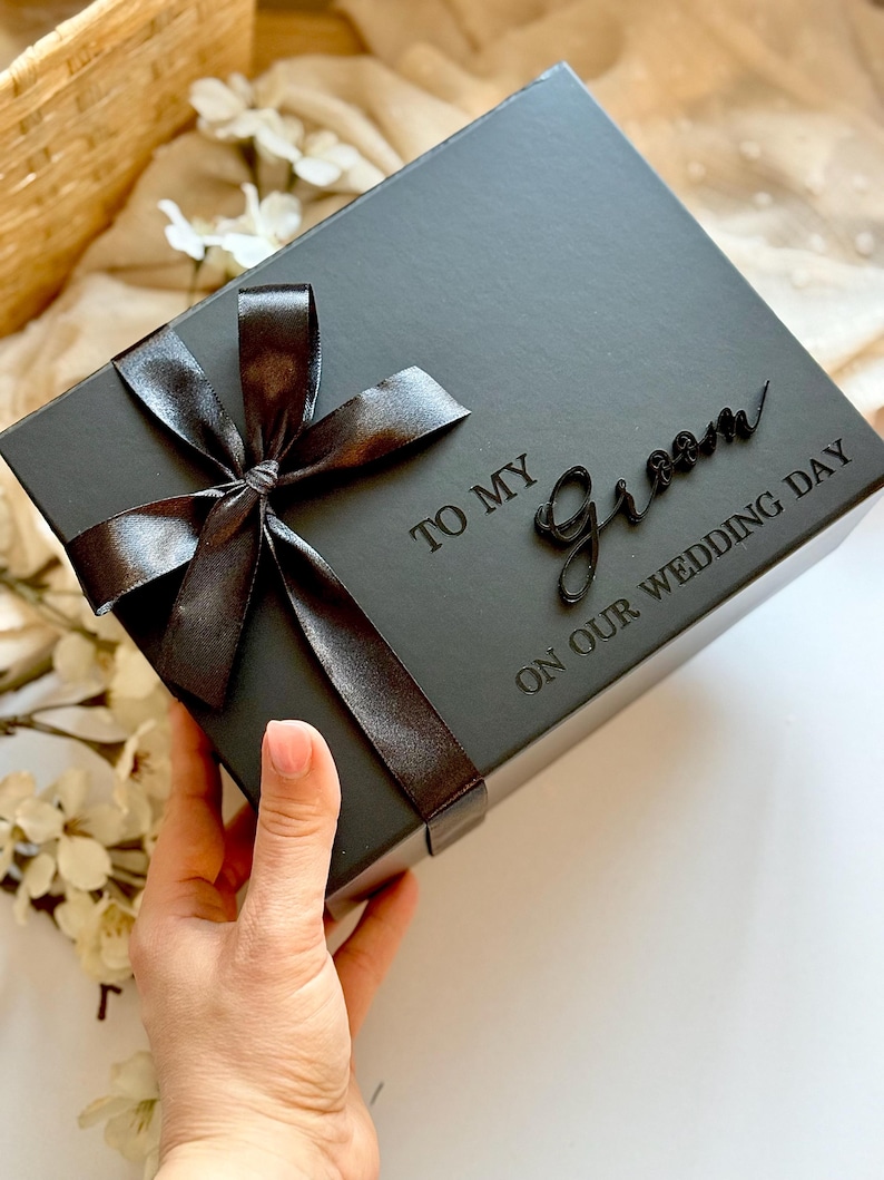 May include: A black gift box tied with a dark brown satin ribbon bow. The box is inscribed with the words "TO MY Groom ON OUR WEDDING DAY" in a script font. The box is held by a person.