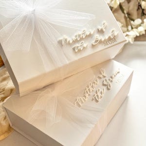 K&ouml;nnte beinhalten: Zwei wei&szlig;e Geschenkboxen, eine &uuml;ber der anderen, sind mit durchsichtigen wei&szlig;en Schleifen verziert. Auf den Boxen stehen die W&ouml;rter "Mother of the Bride" und "Mother of the Groom" in Schreibschrift. Das Wort "MUM" ist ebenfalls auf den Boxen aufgedruckt.