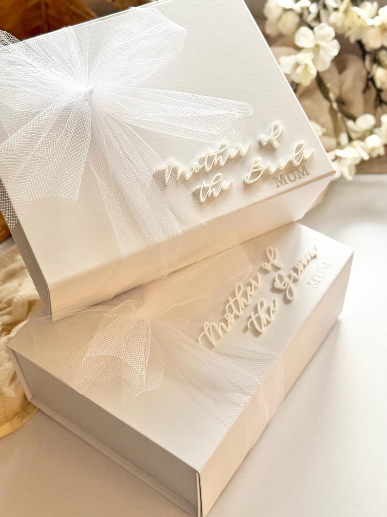 K&ouml;nnte beinhalten: Zwei wei&szlig;e Geschenkboxen, eine &uuml;ber der anderen, sind mit durchsichtigen wei&szlig;en Schleifen verziert. Auf den Boxen stehen die W&ouml;rter "Mother of the Bride" und "Mother of the Groom" in Schreibschrift. Das Wort "MUM" ist ebenfalls auf den Boxen aufgedruckt.
