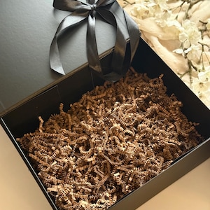 May include: A black gift box with a black satin ribbon tied in a bow. The box is open, showing brown crinkle paper. The box is on a neutral background with white flowers.