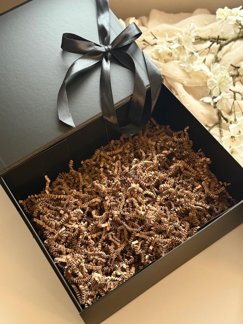 May include: A black gift box with a black satin ribbon tied in a bow. The box is open, showing brown crinkle paper. The box is on a neutral background with white flowers.