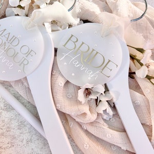 May include: Two white wooden hangers with round, white labels. The label on the left hanger says "Maid of Honour" and the label on the right hanger says "Bride Hannah".