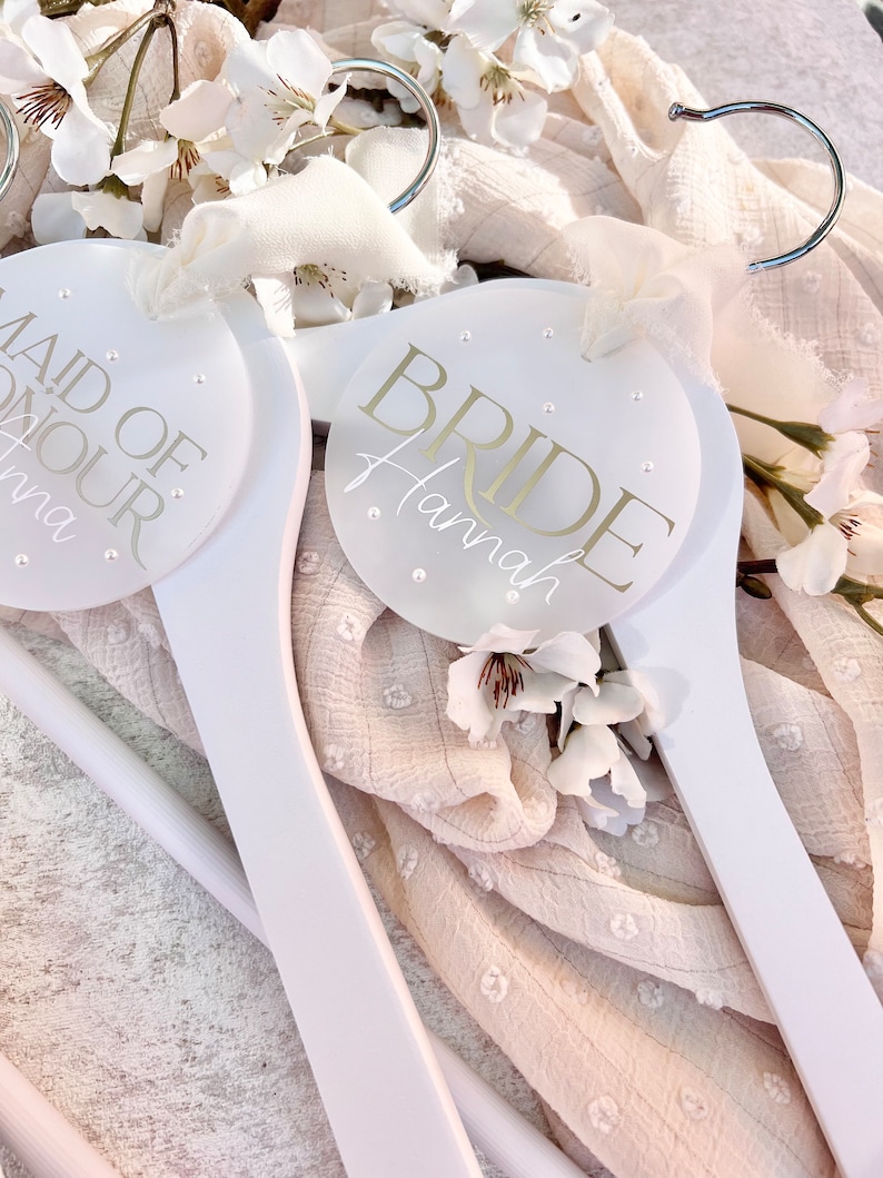 May include: Two white wooden hangers with round, white labels. The label on the left hanger says "Maid of Honour" and the label on the right hanger says "Bride Hannah".