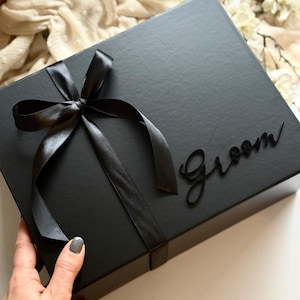May include: A black gift box tied with a black satin ribbon and bow. The word "Groom" is written in a cursive font on the box. The box is rectangular and has a matte finish.