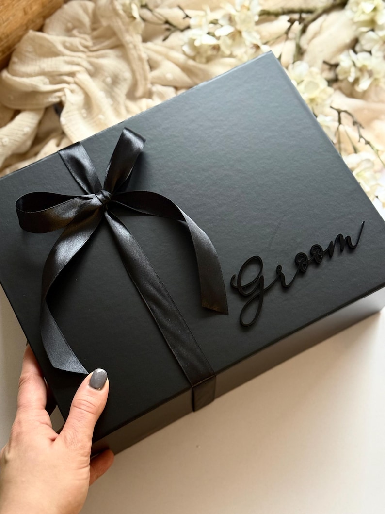 May include: A black gift box tied with a black satin ribbon and bow. The word "Groom" is written in a cursive font on the box. The box is rectangular and has a matte finish.