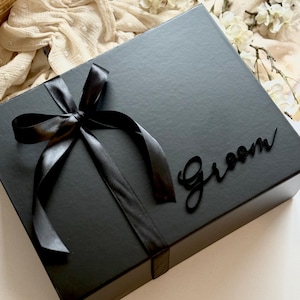 May include: A black gift box tied with a black satin ribbon and bow. The word "Groom" is written on the box in a cursive font. The box is on a white surface.