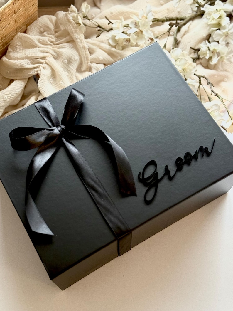 May include: A black gift box tied with a black satin ribbon and bow. The word "Groom" is written on the box in a cursive font. The box is on a white surface.