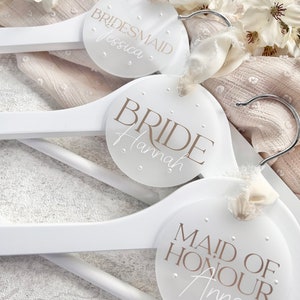 May include: Three white wooden hangers with round, white labels. The labels have the words "Bride Hannah", "Maid of Honour Anna", and "Bridesmaid Jessica" written in brown text.