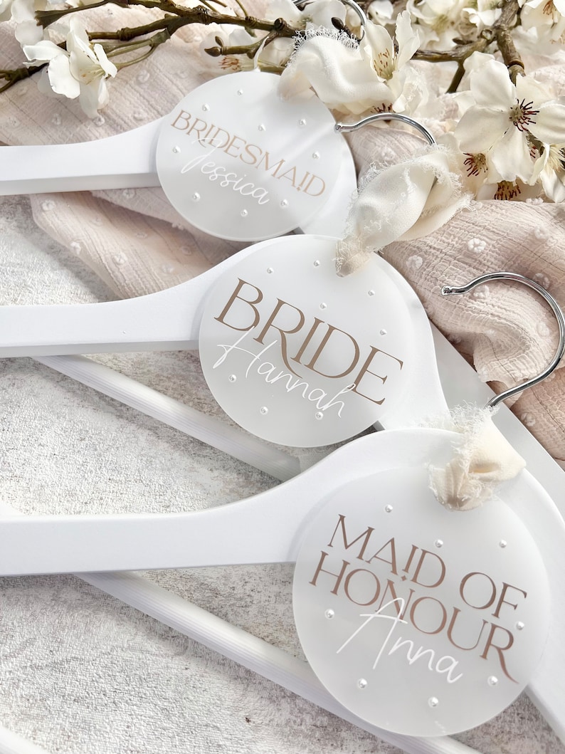 May include: Three white wooden hangers with round, white labels. The labels have the words "Bride Hannah", "Maid of Honour Anna", and "Bridesmaid Jessica" written in brown text.