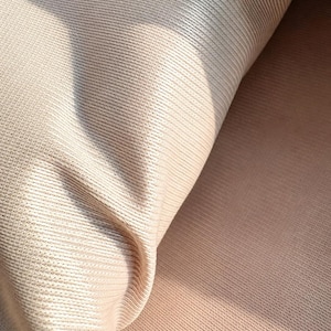 May include: A light beige fabric with a fine, vertical ribbed texture. The material is folded over, revealing a smooth, consistent knit pattern across the surface. The light creates subtle highlights and shadows along the folds of the textile.