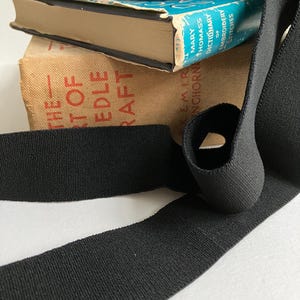 May include: Black elastic band, also known as a fold over elastic, on top of a stack of books. The top book has the title "Mary Thomas's Dictionary of Embroidery Stitches".