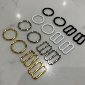 Op de afbeelding: Assortiment van metalen bh-bandverstellers in goud, zilver en zwart. De verstellers zijn in verschillende maten en stijlen, waaronder enkele ringen, dubbele ringen en haak- en oogsluitingen.