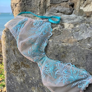 May include: A beige bra with turquoise floral embroidery and a turquoise strap. The bra is made of a sheer material and is resting on a weathered stone wall, with a body of water in the background.