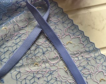 May include: Close-up of blue lace fabric with a floral design and a matching blue ribbon. The lace features a delicate pattern with pink and gold accents. The ribbon is a solid color and has a textured appearance. Purple flowers are in the background.