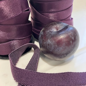 May include: Close-up of spools of deep purple ribbon with a textured edge, and a single plum. The ribbon is tightly wound, and a length of ribbon is unraveled. The plum is a dark purple color.