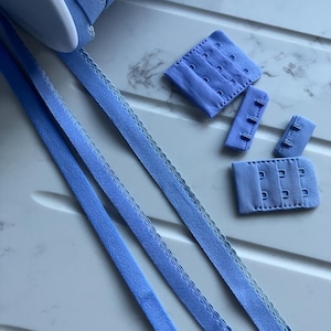 May include: A spool of light blue ribbon with a scalloped edge, along with several light blue bra closure sets. The closures include hook and eye sets in various sizes. The items are arranged on a white surface.