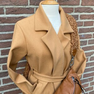 May include: A camel-coloured coat with a belt and a brown leather fanny pack with a fluffy strap.