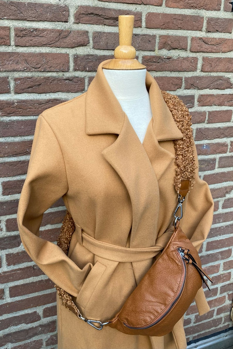 May include: A camel-coloured coat with a belt and a brown leather fanny pack with a fluffy strap.