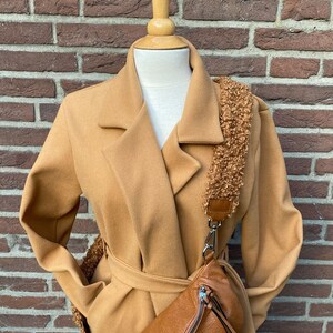 May include: A camel-coloured coat with a belt and a brown leather fanny pack with a fluffy strap.