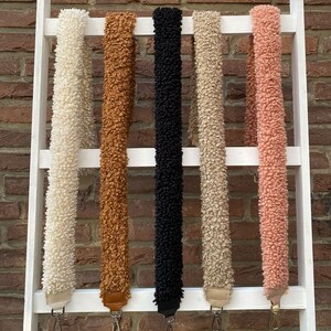 May include: Five different coloured fuzzy straps hanging on a white ladder. The straps are white, brown, black, beige, and pink.