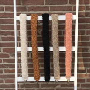 May include: Five fuzzy fabric straps in different colours hanging on a white ladder. The straps are white, brown, black, beige, and pink.