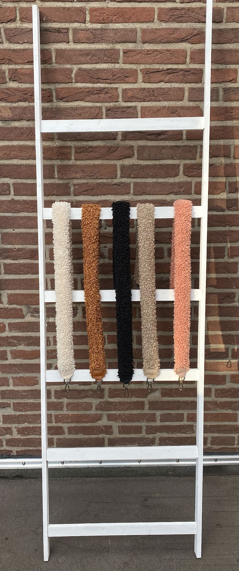May include: Five fuzzy fabric straps in different colours hanging on a white ladder. The straps are white, brown, black, beige, and pink.
