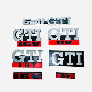 Peut inclure: Ensemble d'emblèmes GTI argentés et noirs, dont "Golf GTI" et divers badges "GTI" avec des accents rouges et noirs. Les badges comportent les mentions "16V", "8V" et "1.8T".