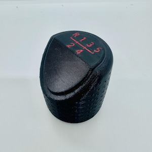 Può includere: Pomello del cambio nero con indicatori del modello del cambio rossi. Gli indicatori includono R, 1, 2, 3, 4 e 5. Il pomello ha una superficie strutturata ed è per un veicolo con cambio manuale.